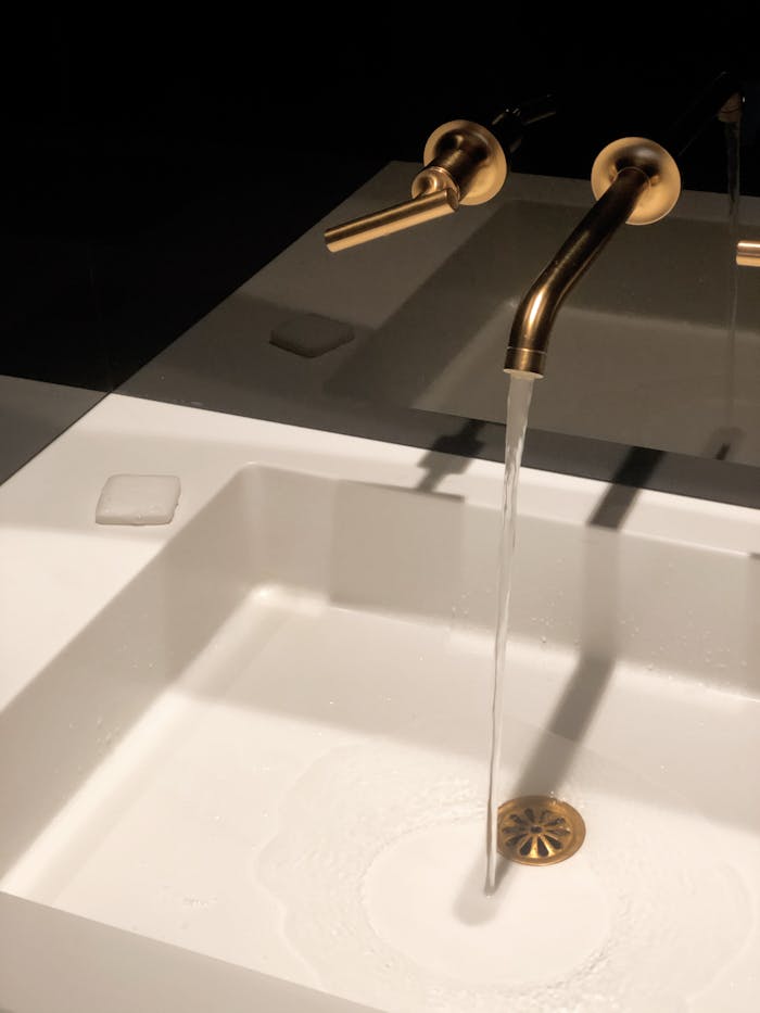 why-choose-us-01 A sleek gold faucet pours water into a modern white washbasin in a dimly lit, elegant bathroom setting.